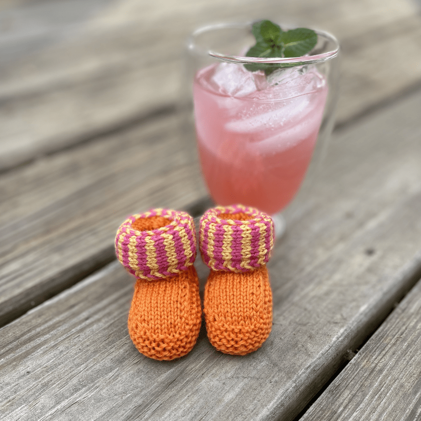 A pair of orange, pink, and yellow knitted baby booties sit on a weathered deck in front of a glass of pink lemonade.