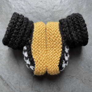 A pair of yellow, black and white knitted booties with a cabled cuff made to look like a car tire sit sole to sole on a gray tile floor.