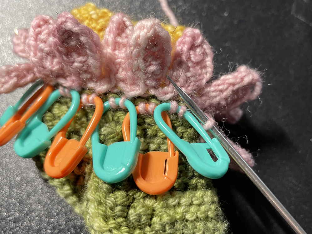 Seven plastic locking stitch markers hold pairs of stitches for petals on a knitted baby bootie made to look like a flower.